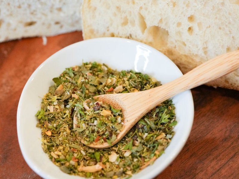 Small white bowl of a green dried herb blend with a wooden spoon, in front of slices of airy sourdough bread on a wooden board.