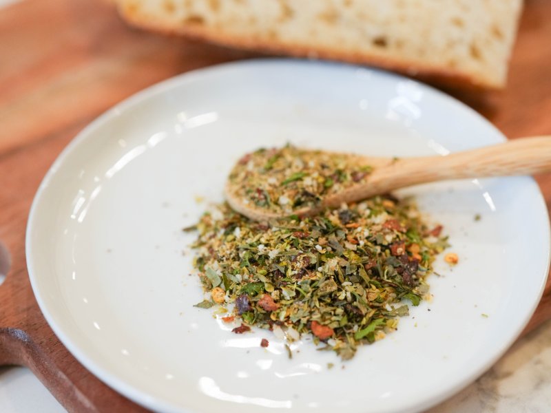 Speckled pile of green herbs and red and black pepper with a wooden spoon on a white plate, beside two slices of sourdough on a wooden board.