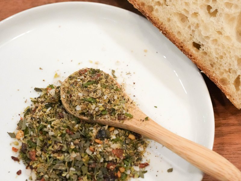 Close-up of a speckled green herb blend with a wooden spoon on a white plate, beside slices of airy sourdough bread.