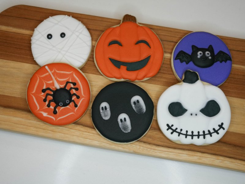 Six decorated cookies on a wooden board: a white mummy with crisscrossed icing wraps and two dot eyes, a smiling orange jack-o’-lantern, a black bat with a happy face on a purple circle, a black spider on an orange cookie with a white web, three little gray ghosts on a black circle, and a white pump