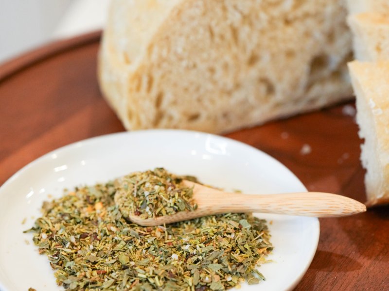 Pile of olive-green dried herbs with a wooden spoon on a white plate, in front of a torn sourdough loaf on a round wooden board.