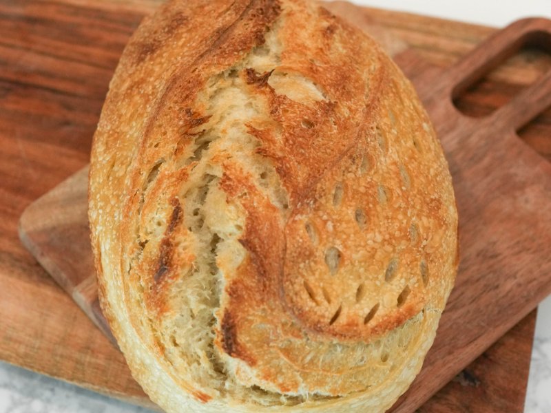 Golden-brown oval Olive Oil & Honey sourdough loaf with a wide curved score and leaf-shaped cuts, on wooden boards over white marble.