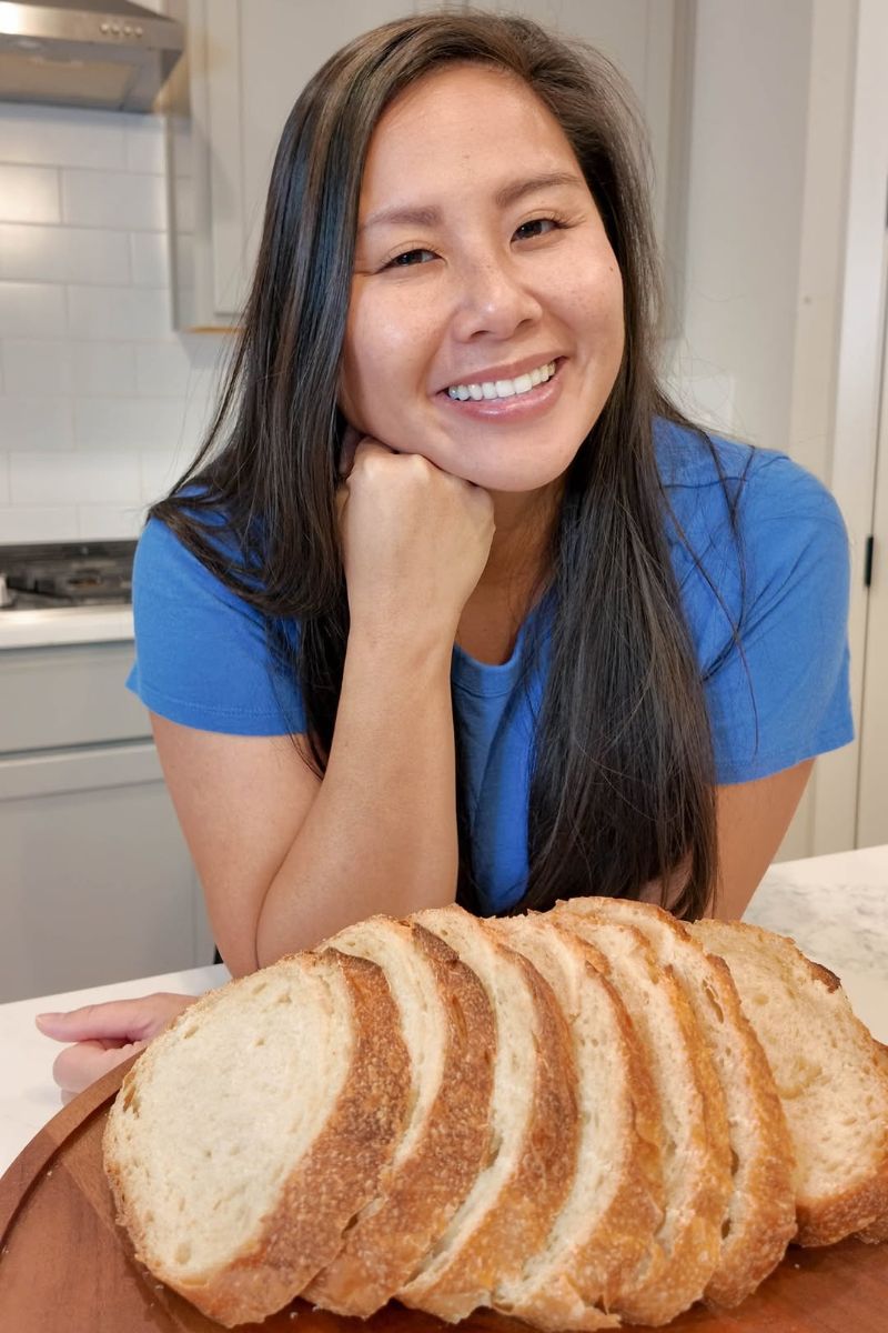 Vy (Mary) smiling in her kitchen, chin on her hand, behind a sliced sourdough loaf on a wooden board.
